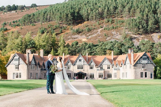 bride and groom standing in front of large country house with Scottish hills surrounding them